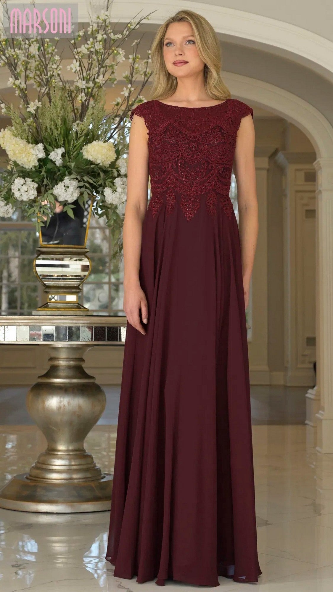 A woman stands indoors in a wine Marsoni M238 long formal dress with an embroidered bodice and flowing chiffon skirt, posed before a large white flower arrangement.
